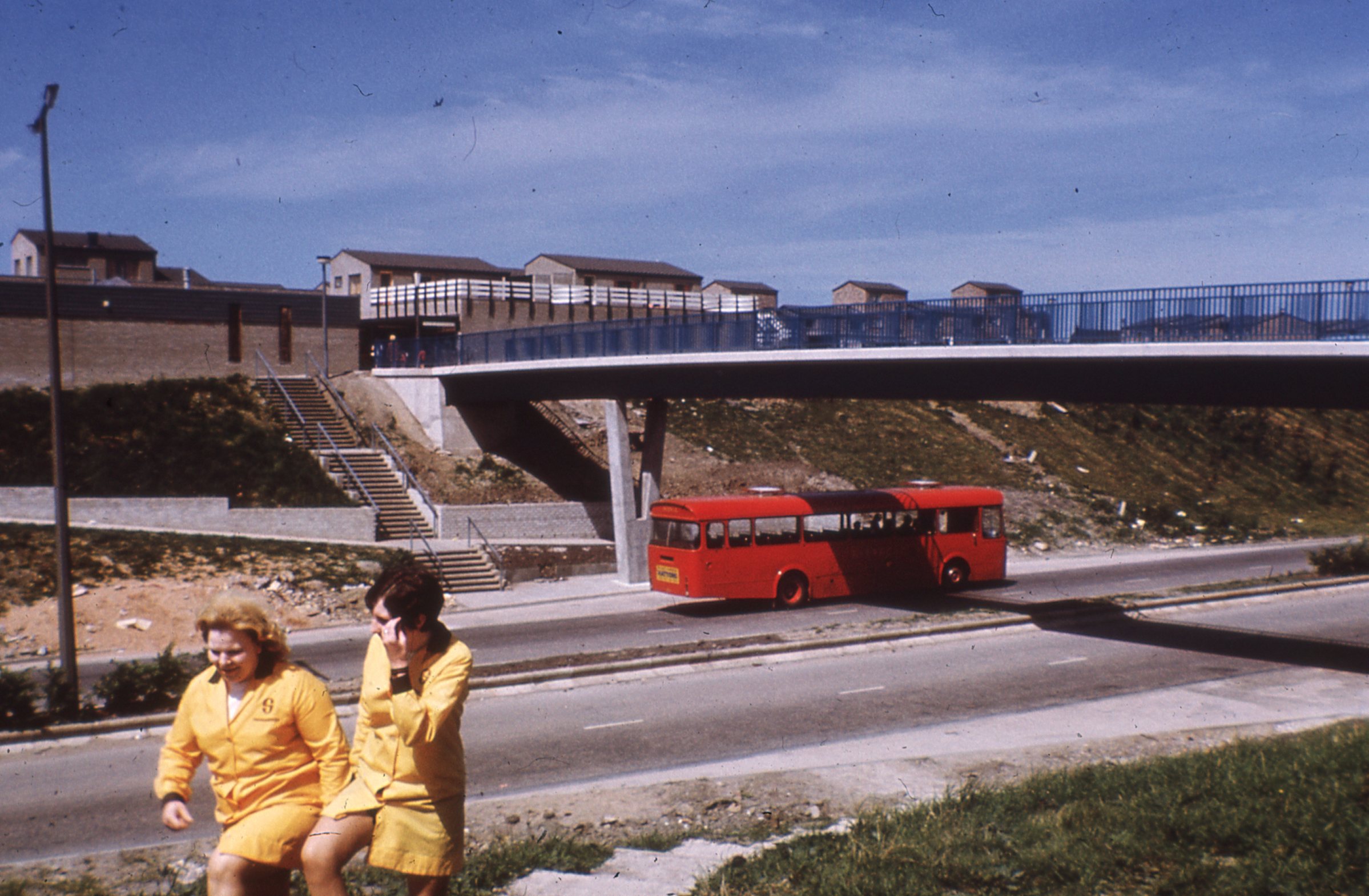 A view looking over a main road towards some 1960s buildings. A red bus is on the road. Two women wearing yellow uniforms walk towards the camera.