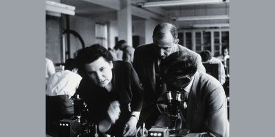 A black and white photograph showing a woman and two men bending over a microscope in a lab.