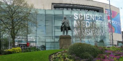 A photo of The National Science and Media Museum in Bradford, with a tree, lawn, plants and a statue in front of it.