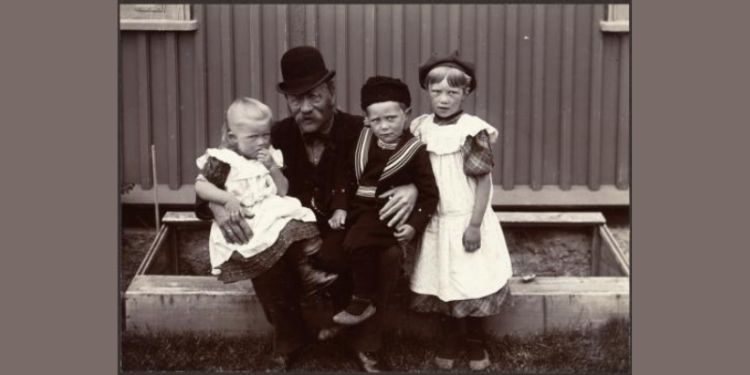 A man with glasses, wearing a hat holding a little girl and little boy on his lap, older girl standing next to him.