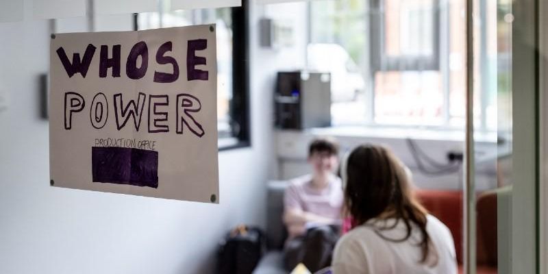 Two people in a room with a sign on the window that reads Whose Power production office.