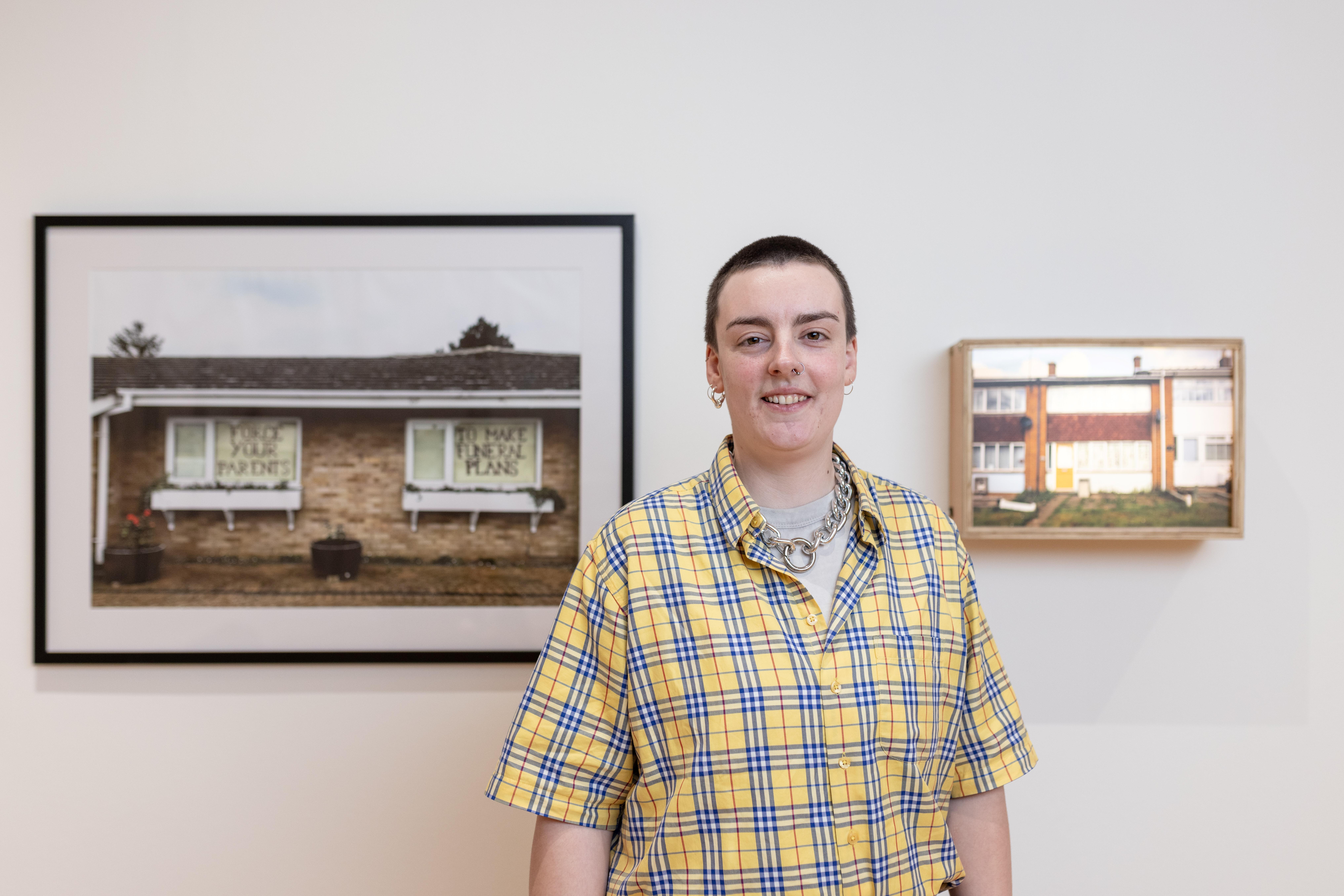 FAHACS student George Storm Fletcher photographed in front of their work featured at Leeds Art Gallery for Leeds Artists Show 2023. George is looking directly into the camera and smiling. To the left and right of George are two pieces of their work.