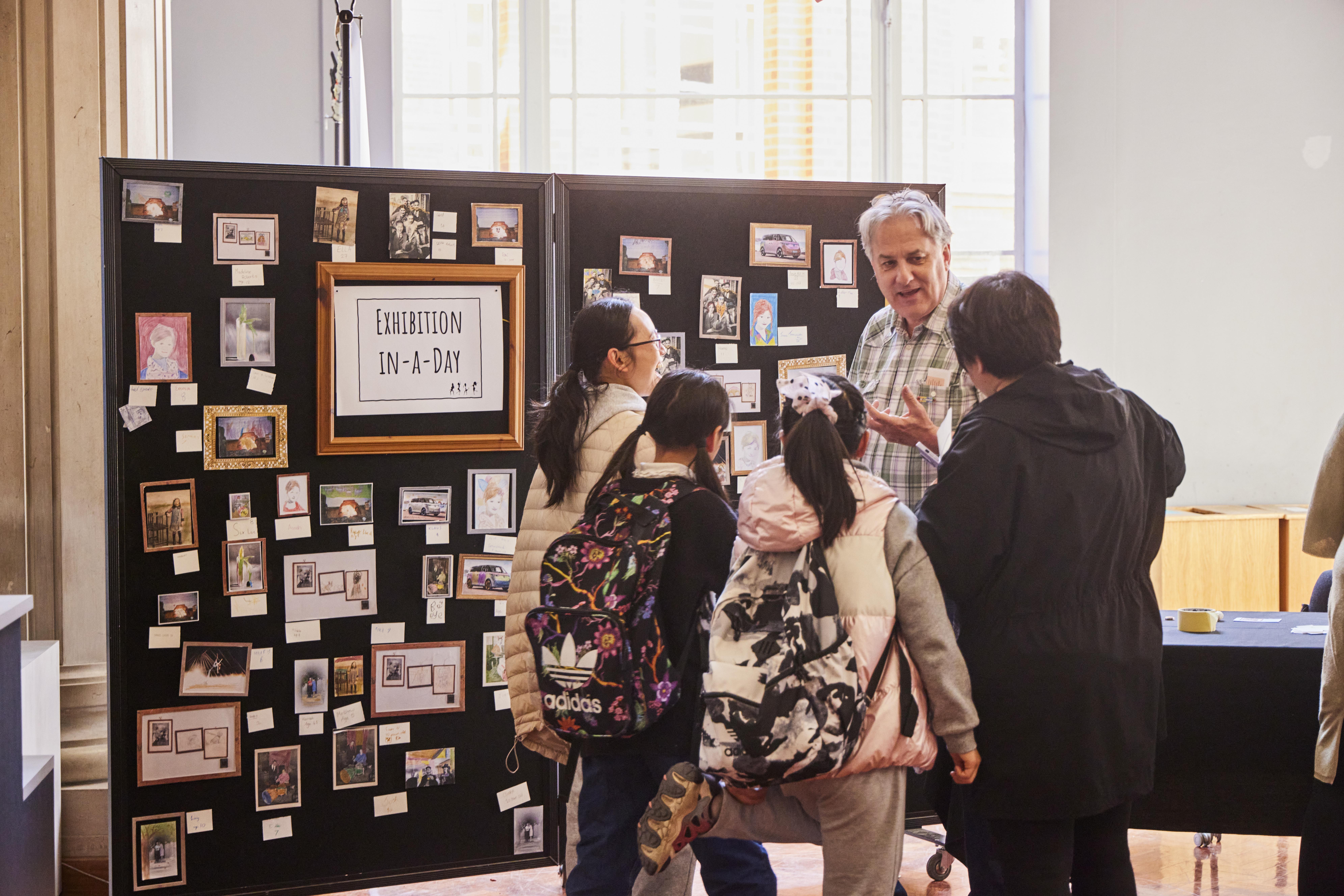 A family looking at a board titled 'Exhibition in a Day', which has photographs pinned all over it.
