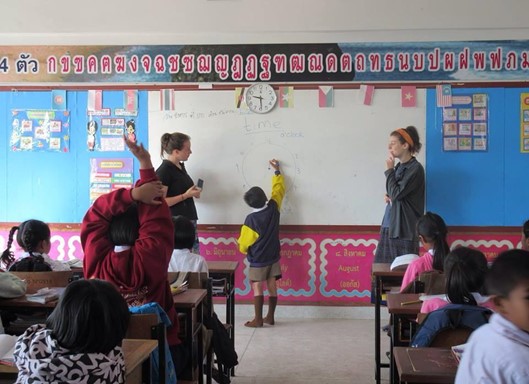 A thai classroom with child writing on a board being supervised by two teachers