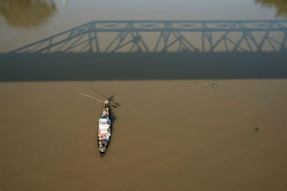 Photograph of a traditional wooden boat on the Surma River passing under Keane Bridge in Sylhet