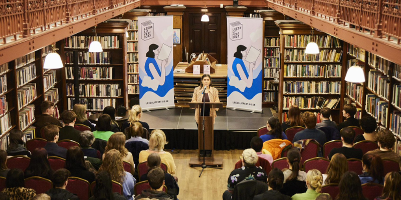 A person behind a podium at Leeds Lit Fest 2023. They're in the Leeds Central Library. A crowd is seated before them as they speak.