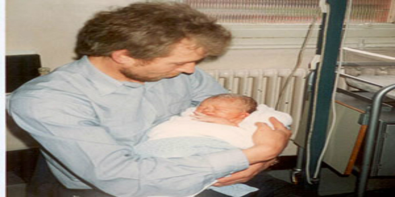 A photo of a man in a blue shirt holding a baby wrapped in a blanket.