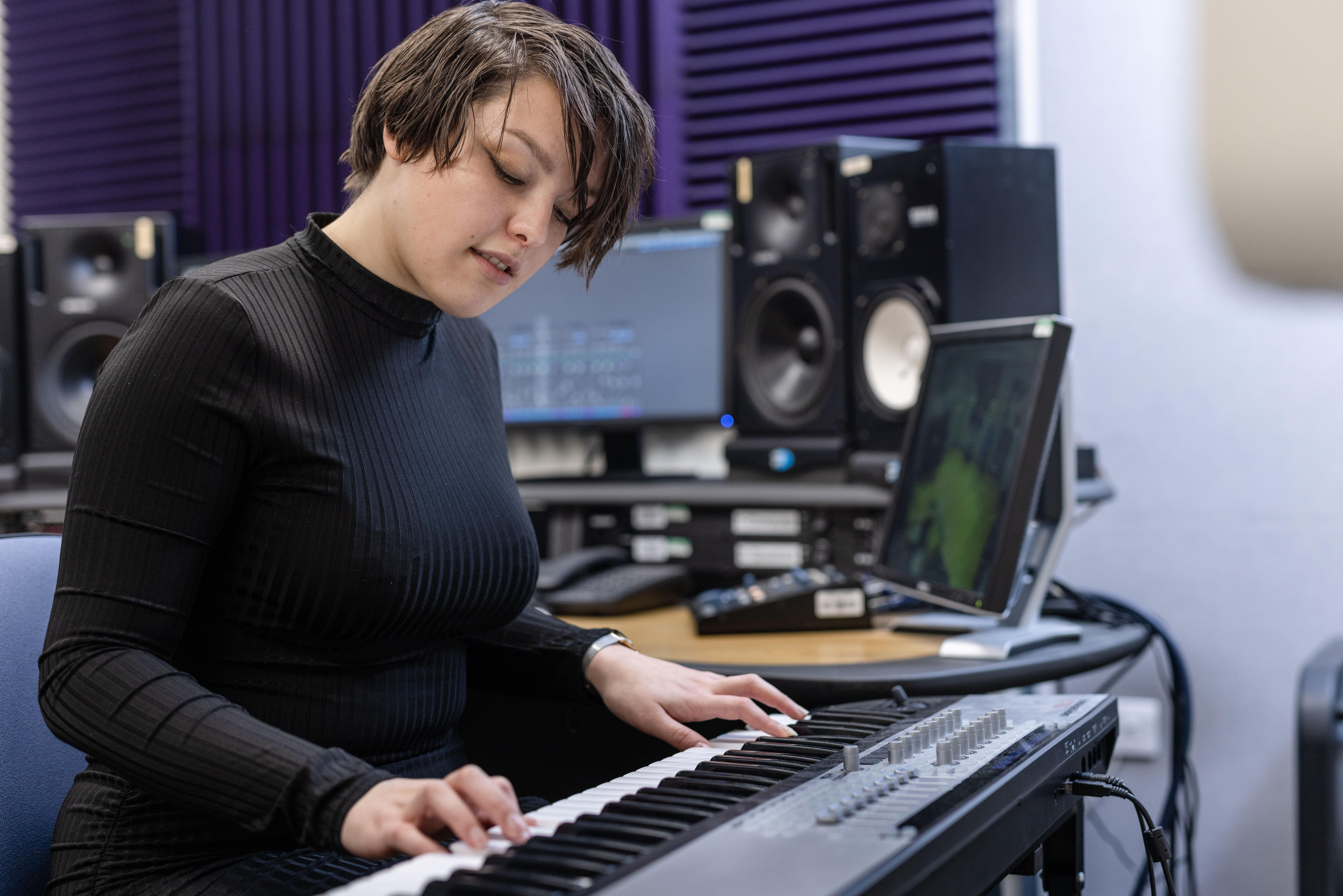 Student practicing on the keyboard in the electronic studios. In the background is a screen, speakers and other electronic equipment.