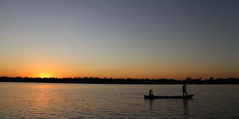 Two people on opposite ends of a canoe with the sun peeking out behind the horizon
