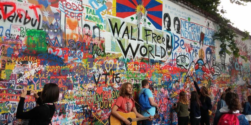 Approx 10 people (incl one playing a guitar) standing in front of the grafitti-covered Lennon Wall looking at the grafitti.