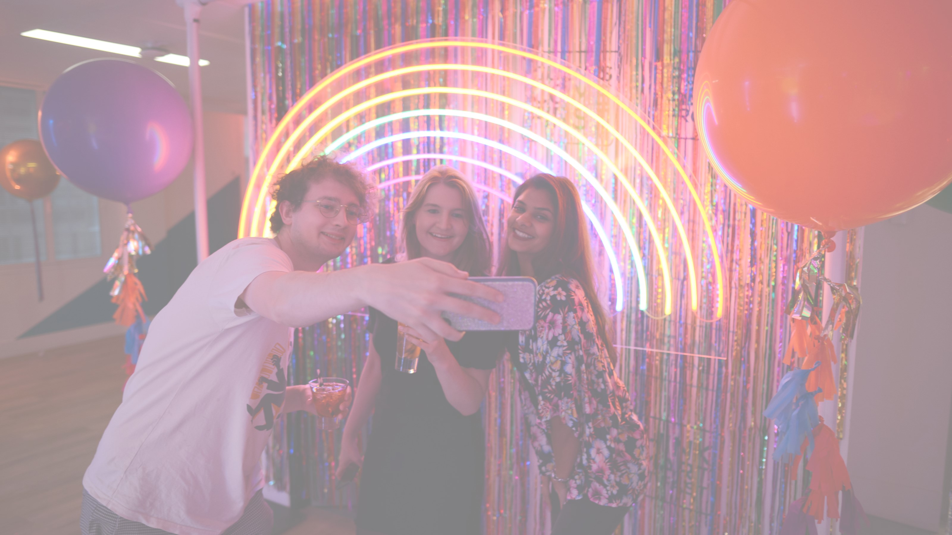Three young people smiling, taking a selfie in front of a neon rainbow light, balloons and streamers.