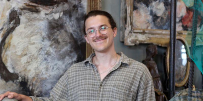 School of Music alumnus Ben Adamson. Ben is in a gallery setting and looks directly at the camera and is smiling. He is wearing a shirt and glasses.