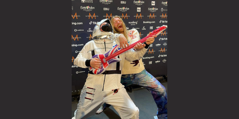 Sam Ryder in front of a backdrop with Eurovision logos on it. He&#039;s with somebody dressed as a spaceman with a blow up guitar.
