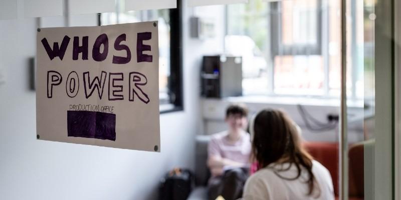 Two people in a room with a sign on the window that reads Whose Power production office.