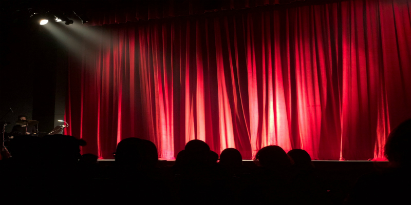 An audience sit in the dark watching a drummer on stage in front of a red curtain.
