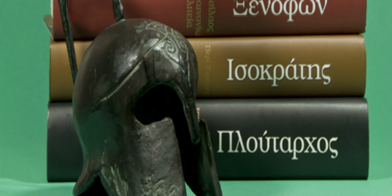 An image of a Greek-style helmet in front of Greek books with a green background.