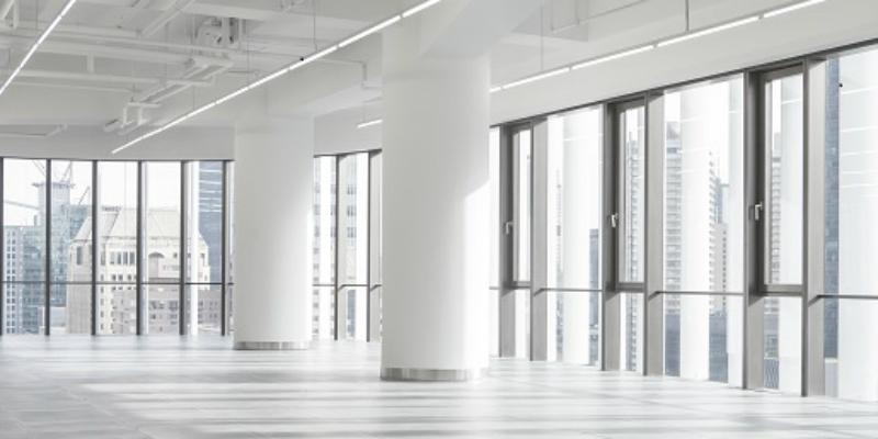 A photo of an empty office with a white floor, ceiling and columns, and floor to ceiling glass windows with high-rise buildings viewable from them.