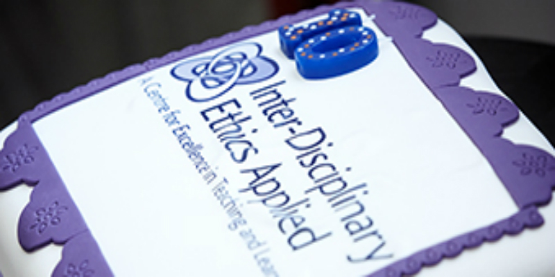 An image of a cake to celebrate the IDEA Centre's 10th anniversary. It is a square cake covered in white icing, with a purple icing trim, and blue candles and writing.