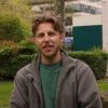 Student Alex Lewis sat outside with bushes, trees and grass in the background. Alex wears a grey fleece with green t shirt and is smiling.