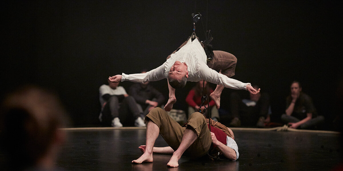 Two actors in historical costume on stage - one suspended horizontally over the other