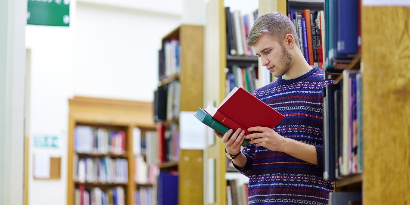 A male student leans against a bookshelf in a library, a book in his hands as he reads it
