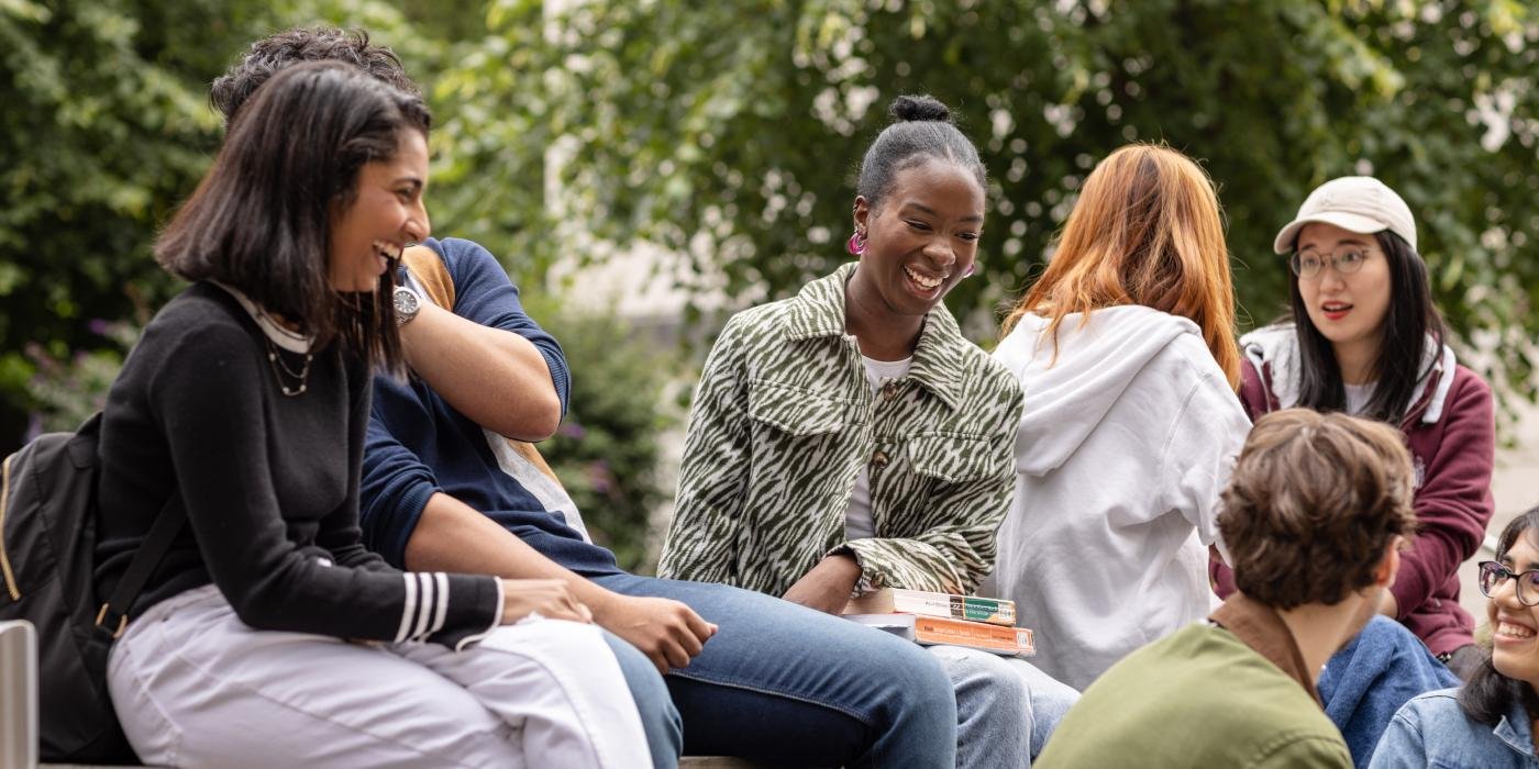 A group of students sit together on campus, they are laughing and joking, with multiple conversations taking place within the group.