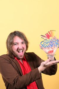 Award winner stood against yellow background, smiling holding award wearing red top and brown jacket