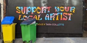 Two wheelie bins in front of a large, landscape black poster with colourful text 'Support your local artist'.