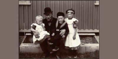 A man with glasses, wearing a hat holding a little girl and little boy on his lap, older girl standing next to him.