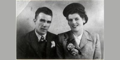 Two young people in formal clothes smile at the camera. They have floral buttonholes and the lady wears a hat.
