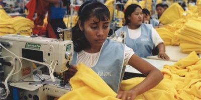 Crowded garment factory with workers operating sewing machines and handling piles of yellow fabric in a high-pressure, mass-production environment typical of sweatshops