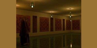 A dark room with Islamic tapestries on the wall and lamps hanging from the ceiling. A veiled woman stands to the left, facing an archway.