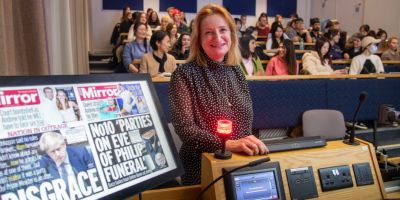 Alison Phillips stands smiling next to digital copies of covers of Mirror newspapers in front of a class full of students.