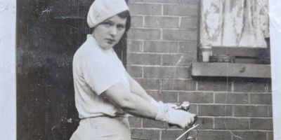 A Black and white photograph of a young woman wearing a beret looking the camera in front of a brick wall. Her hands are on bicycle handles