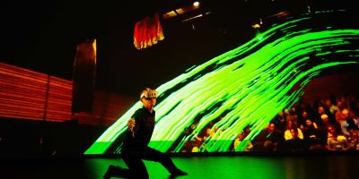 A dancing in a black suit kneeling on a stage with a VR headset.