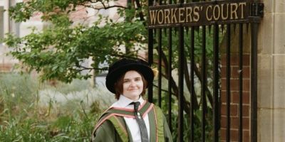 A figure wearing Leeds PhD robes of green and orange smiles to the camera