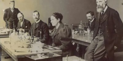 Men and women from the Victorian era stand around laboratory benches