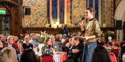 Heather Peak in the Civic Dining Room, Rochdale Town Hall. Heather is speaking into a microphone to an audience of people sitting round tables.