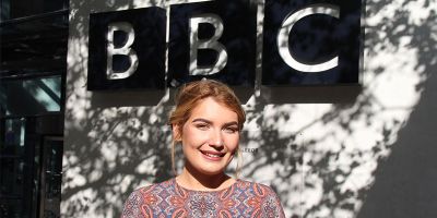 Jessica Forrester stands outside BBC Radio Leeds smiling in the sunshine with the BBC logo on the building behind her head with shadows cast on the white walls by a tree.