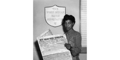 Black and white image of a Una Marson stood against a wall reading a copy of the West Indian Radio Newspaper, during WWII