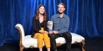 Charlotte Field and Hugh Clegg sit on a white chaise longue, holding their glass award together, in front of a blue velvet curtain.