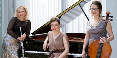 Three musicians in front of a grand piano. Left: Helen Vidovich holding a flute, leaning onto the side of the piano. Middle: Olga Stezhko, sitting in front of the piano and leaning back onto it with one elbow. Right: Val Welbanks standing, holding a cello