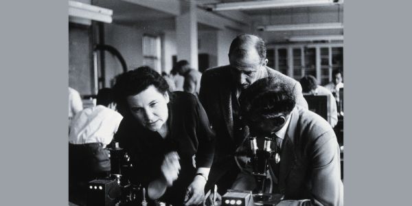 A black and white photograph showing a woman and two men bending over a microscope in a lab.