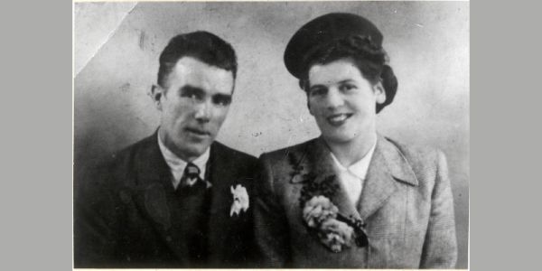Two young people in formal clothes smile at the camera. They have floral buttonholes and the lady wears a hat.