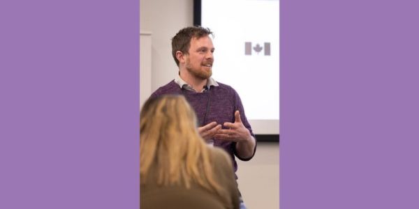 A man with a short beard wearing a purple jumper standing in front of a whiteboard. The Canadian flag is visible on the whiteboard.