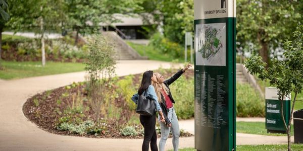 Two female students on campus. They are looking up at and pointing to a large campus map as they figure out where they need to go.