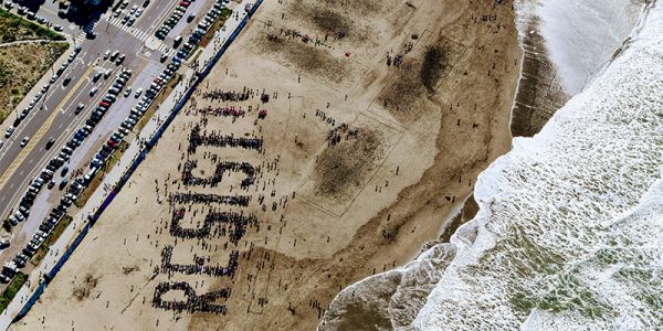 A birs-eye view of people spelling out the word 'resist!' on a beach, with waves lapping the shore.