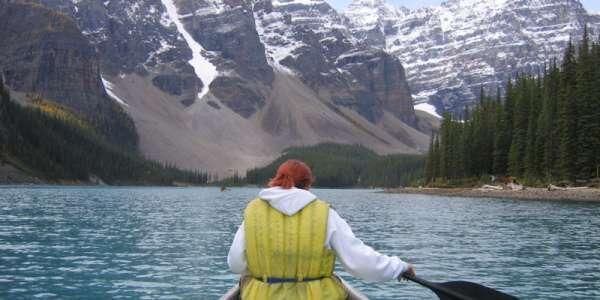 A person kayaking in a yellow vest, their back is to the camera and ahead of them is a mountain range.