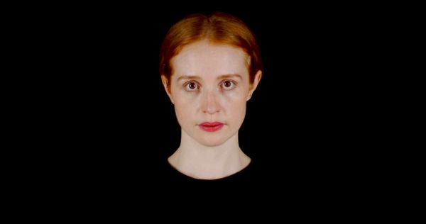 A young woman with red hair and light skin is staring directly at the camera against a black background. She is wearing a black top.