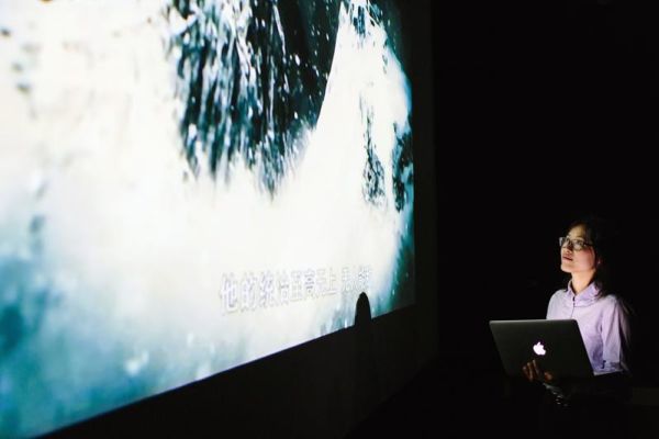 A student holding a laptop watches an abstract film projection on a large screen in a dark room. The projection includes foreign language subtitles, and the student is focused on the screen.